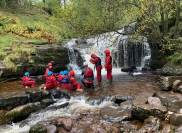 A group of teenagers wearing red waterproof clothing standing and sitting in a river with a waterfall in the background.