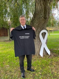 PFCC Roger Hirst standing outside holding up a T-Shirt that promotes White Ribbon Day with a large White Ribbon logo board next to him.