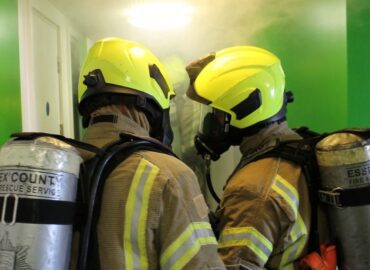 Two firefighters wearing breathing apparatus inside the corridor of a building.