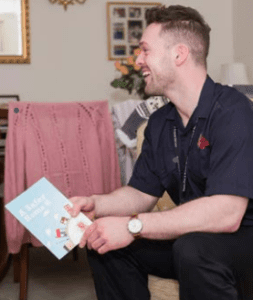 An Essex Fire and Rescue Service employee is sitting on the sofa during a safe and well visit offering advice. He is wearing a blue uniform and is smiling and holding leaflets containing safety information.