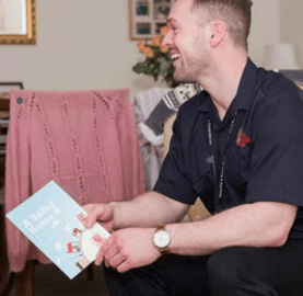 An Essex Fire and Rescue Service employee is sitting on the sofa during a safe and well visit offering advice. He is wearing a blue uniform and is smiling and holding leaflets containing safety information.