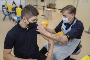 Firefighter giving a man a Covid 19 vaccination at a vaccination centre. They are both wearing face coverings.