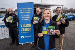 Four people standing in front of a blue banner which says Vision Zero. They are smiling and holding leaflets about road safety.