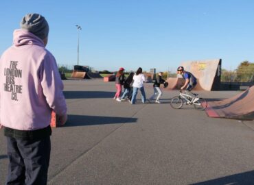A group of teenagers are playing in a skateboard/BMX park. One is riding a BMX bike. A man wearing a London Bus Theatre Co hooded top is encouraging them in the foreground.