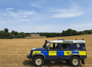 In this image there is an Essex Police branded Land Rover 4 x4 vehicle parked in a stubble field with Audley End House in the background.