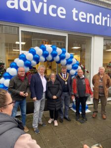 Volunteers and special guests including local MP Giles Watling at the opening of the new centre. They are standing in front of the shop underneath a white and blue balloon arch.