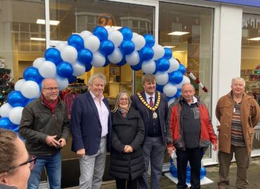 Volunteers and special guests including local MP Giles Watling at the opening of the new centre. They are standing in front of the shop underneath a white and blue balloon arch.